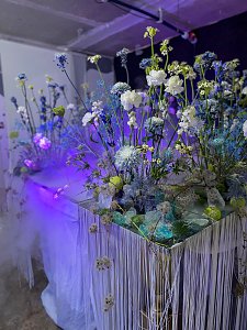 Üppige florale Dekorationen aus blau-weißen Blumen, die unter kühlem, lila-blauem Licht auf einem mit Fransen verzierten Tisch arrangiert sind.