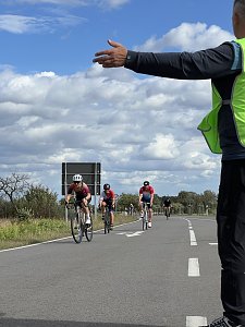 Radstrecke beim Triathlon. Im Vordergrund weist ein Streckenposten mit ausgestrecktem Arm den Weg. Dahinter fahren mehrere Radfahrer mit Helmen auf einer asphaltierten Straße. Im Hintergrund ist ein bewölkter Himmel zu sehen.
