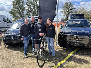 Vier Mitarbeiter des Autohauses Peter stehen für ein Gruppenbild auf einer Wiese zwischen zwei schwarzen Mercedes-Fahrzeugen. Der Mann in der Mitte trägt eine Medaille und hält ein Rennrad. Im Hintergrund weht eine große schwarze Fahne.