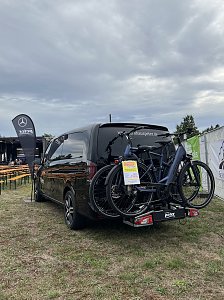 Eine schwarze Mercedes-Benz V-Klasse mit Fahrradträger, auf dem zwei E-Bikes befestigt sind. Im Hintergrund das Festgelände.