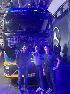 Drei M&auml;nner stehen l&auml;chelnd vor der Front eines gro&szlig;en Mercedes-Benz Lkws. Das Fahrzeug ist dunkel lackiert, hat gelbe Akzente unten und eine Weltkarte aufgedruckt. Die gesamte Szene auf der Messe ist in intensives, blaues Licht getaucht.