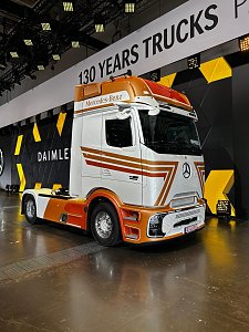 Wei&szlig;-orangefarbener Mercedes Lkw von vorne rechts auf einer Messe. Am Fahrerhaus kleben gold-rote Streifen. Im Hintergrund h&auml;ngt ein Banner mit der Aufschrift 130 YEARS TRUCKS. Der Lkw steht auf einem gl&auml;nzenden, spiegelnden Messeboden.