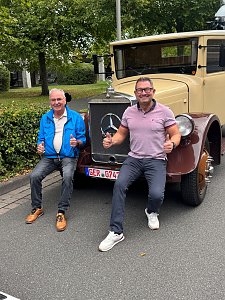 Zwei lachende M&auml;nner sitzen auf den Kotfl&uuml;geln eines beigefarbenen Oldtimer-Lkws mit braunen Radl&auml;ufen. Beide zeigen den Daumen nach oben. Der Mann links tr&auml;gt eine blaue Jacke, der rechte ein rosa Poloshirt. Im Hintergrund stehen gr&uuml;ne B&auml;ume.