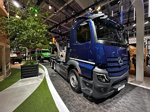 Dunkelblauer Mercedes-Benz Lkw von vorne rechts auf einem Messestand. Im Hintergrund links steht ein gr&uuml;nes Nutzfahrzeug neben einem gro&szlig;en, echten Baum im Pflanzk&uuml;bel. Der Boden hat wei&szlig;e Linien und gr&uuml;ne Kunstrasen-Fl&auml;chen.