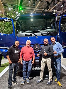 Vier l&auml;chelnde M&auml;nner posieren eng nebeneinander vor einem gro&szlig;en, blauen Mercedes-Benz Lkw auf einer Messe. Die beiden T&uuml;ren des Lastwagens stehen weit offen.