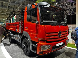 Roter Mercedes-Benz Kipper-Lkw von vorne rechts auf einem Messestand. Das Fahrzeug hat ein kurzes Fahrerhaus, schwarze Felgen und eine rote Ladefl&auml;che hinten. Personen laufen im Hintergrund vorbei. Der Lkw steht auf grauem Teppich.