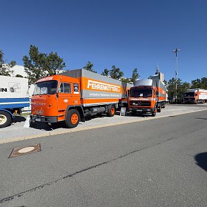 Orangefarbener Oldtimer-Lkw von vorne links auf einem Parkplatz. Er hat eine graue Plane mit der Aufschrift FERNVERKEHR. Dahinter stehen zwei weitere, baugleiche Lastwagen in orange.