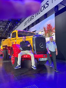 Zwei M&auml;nner posieren mit einem gelb-roten Mercedes-Benz Oldtimer-Lkw auf einer Messe. Ein Mann sitzt l&auml;chelnd auf der roten Sto&szlig;stange vor dem gro&szlig;en K&uuml;hlergrill. Der andere steht rechts daneben. Oben h&auml;ngt ein Banner mit 130 YEARS TRUCKS.