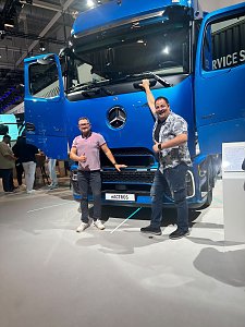 Zwei lachende M&auml;nner posieren vor einem gro&szlig;en, blauen Mercedes-Benz Lkw auf einer Messe. Beide T&uuml;ren des Lastwagens stehen weit offen.