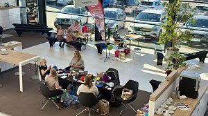 Besucherinnen sitzen an einem Tisch im Autohaus und besch&auml;ftigen sich mit bunten H&auml;kelarbeiten. Im Hintergrund basteln Kinder.