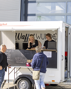 Zwei Mitarbeiterinnen von WaffelWunder bedienen G&auml;ste an ihrem Verkaufswagen auf dem Au&szlig;engel&auml;nde der Autowelt Peter.