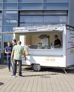 G&auml;ste unterhalten sich vor dem Verkaufswagen des Caf&eacute; Central auf dem sonnigen Au&szlig;engel&auml;nde der Autowelt Peter.
